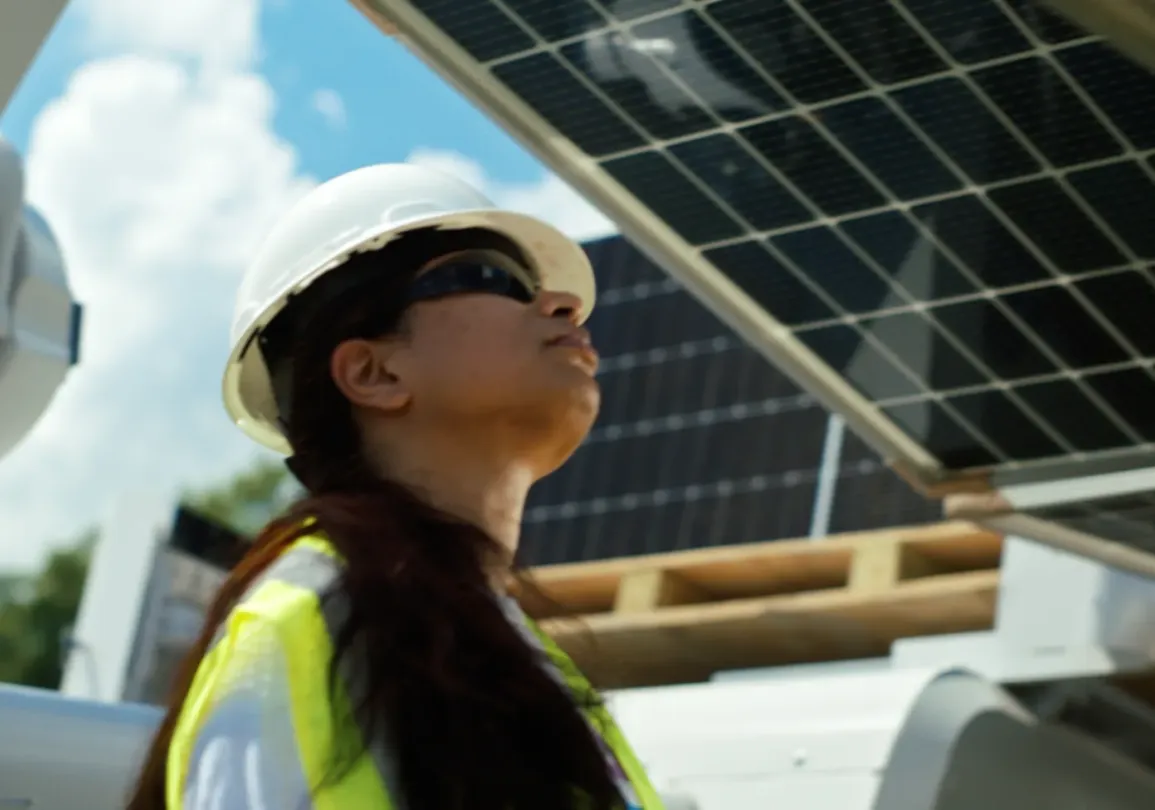 A person wearing a hard hat and safety vest inspects solar panels under a blue sky.
