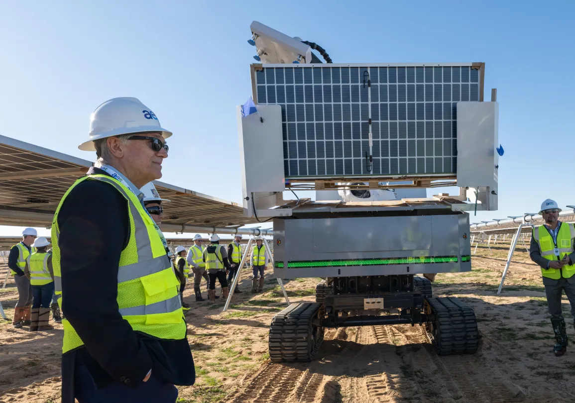Group of workers wearing safety vests and hard hats standing near a solar panel installation and a robotic vehicle at a solar farm on a sunny day.